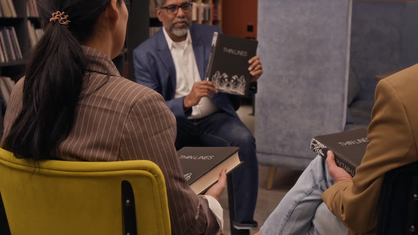 Low angle shot of biracial male author talking about his new book to audience, holding copies of his books, while sitting in library