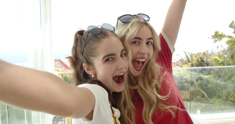 Two female friends holding smartphone for selfie, wearing sunglasses and lip-syncing in sunroom. Casual, friendship, lifestyle, candid, vibrant, dynamic, outdoor