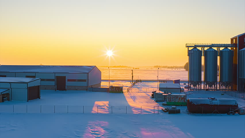 Farm buildings with grain silos on snowy field during golden hour winter morning in aerial hyperlapse