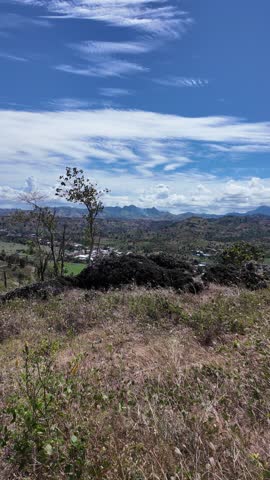 Scenic highland landscape view with dark volcanic rocks and dry grass in the foreground, overlooking a sprawling rural valley, village, and rugged mountains under a bright blue sky.