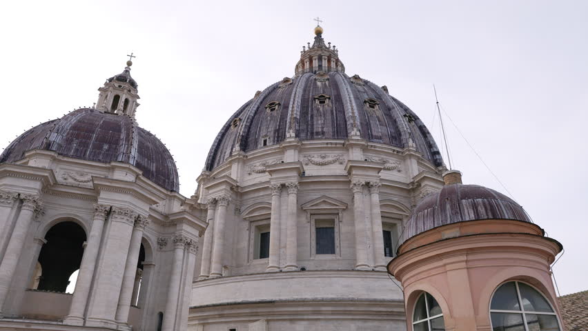 Low angle view of St. Peter’s Basilica dome in Vatican City with monumental travertine architectural details against a bright sky. Iconic symbol of Rome, religion, art and travel.