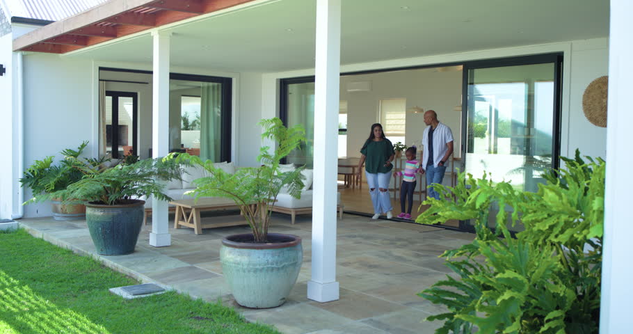 Diverse family of three stepping through sliding doors onto patio, holding hands and surveying yard. Homeowners, daughter, planters, ferns, greenery, sunlit, exterior
