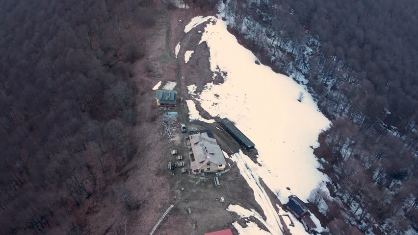 Aerial drone tilt up from Mazalat Hut to surrounding mountain peaks in Stara Planina, Bulgaria. Early spring landscape with patchy snow and forested ridges under soft evening light.