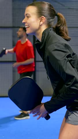 Side view of a focused young woman playing pickleball, smiling as she hits the yellow ball with her paddle during a competitive match with her male partner on an indoor blue court