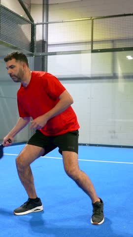 Athletic man in red shirt intensely playing pickleball, hitting the yellow ball with a paddle on a vibrant blue indoor court during a competitive match, showcasing concentration and skill