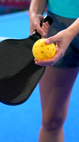 Close up of a female athlete holding a pickleball paddle and a yellow ball, preparing to serve on a bright blue court during a match, representing a healthy and active sport lifestyle
