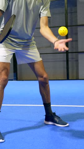Close up of a man serving a yellow pickleball with a paddle on a blue court. The athlete is hitting the ball in slow motion during a match, showcasing the technique and movement of the sport