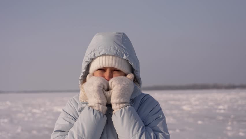 Frigid air surrounds a person bundled in a coat and hat. They press mittens to their face, seeking comfort from the biting wind across a wide, shimmering icy field in broad daylight.