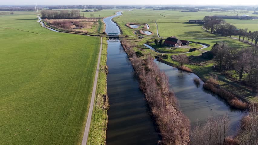 Aerial view of a Dutch canal system with a small dam, lush green farmland, and a traditional brick farmhouse. The scene captures the intricate water management and peaceful rural life in spring.