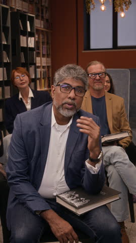 Vertical shot of middle-aged biracial man talking to speaker while sitting among attendees of book presentation