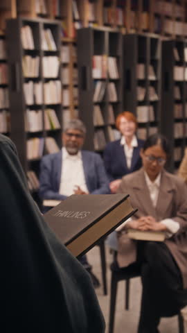 Vertical low angle over the shoulder shot of author holding new book while presenting to group in library