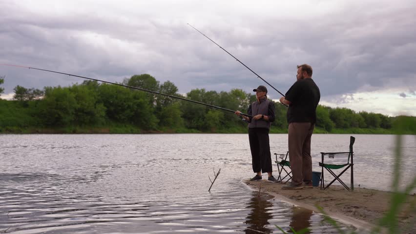 Man and woman spending quality time together, fishing with rods from the sandy bank of a river on a cloudy day, enjoying their outdoor hobby, leisure, and a quiet weekend retreat