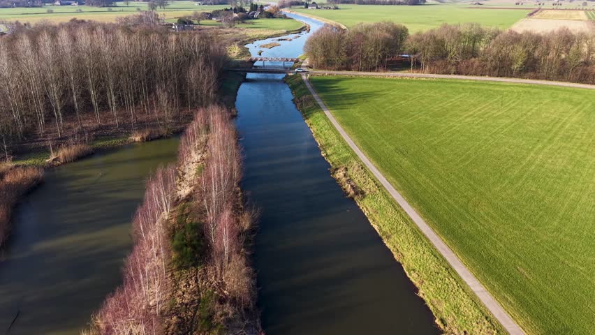 An aerial drone view of a calm canal cutting through a vibrant green rural landscape, featuring a small bridge, a parallel cycling path, and late winter trees under soft sunlight.