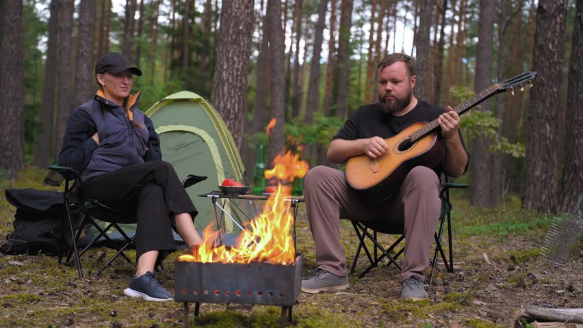 A young couple enjoys a peaceful evening in the woods, where a man plays an acoustic guitar and sings songs to his girlfriend while sitting by a warm, crackling fire near their tent