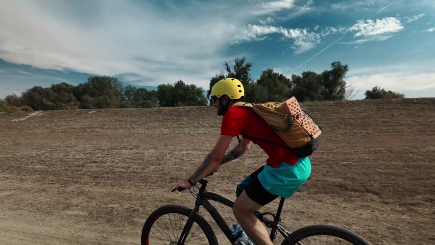 Side view of a man cycling on a dirt road through dry countryside under a blue summer sky. Gravel riding, endurance training and active outdoor lifestyle concept