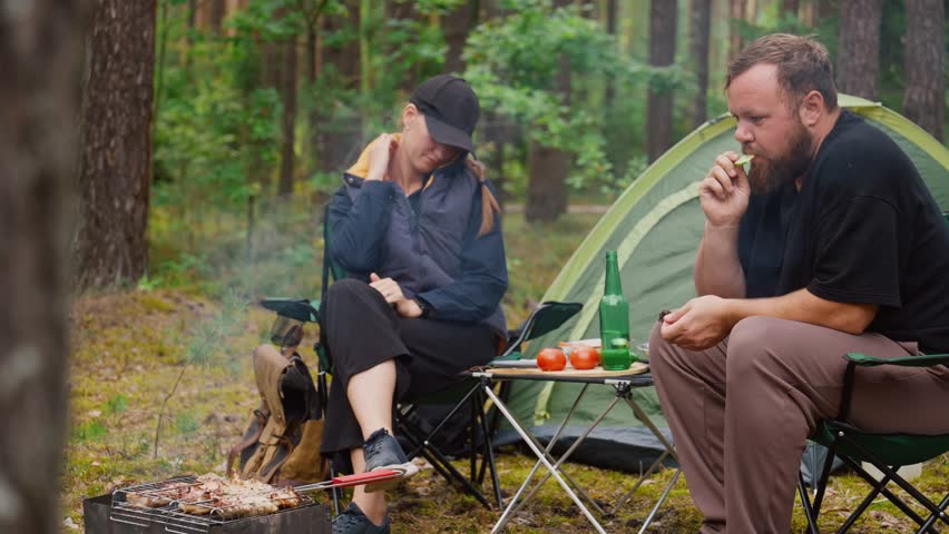 Happy young couple sitting on camping chairs in woods, talking and laughing together, grilling meat on barbecue picnic during their trip to the fresh air and drinking beer