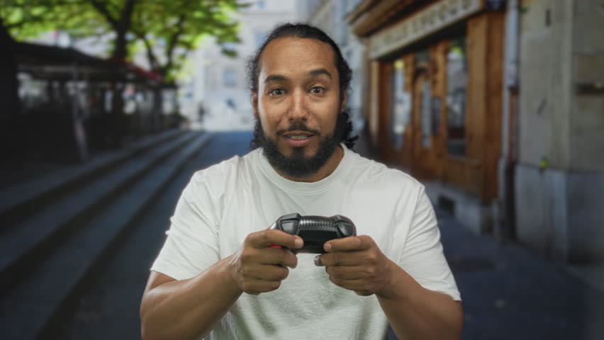Man holding black and red game controller with both hands and raising his right fist while looking at camera on a city street lined with shops and steps; excitement.
