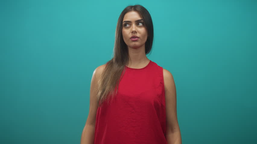 Woman with hand on chest, open mouth and wide eyes, wearing a red sleeveless shirt against a teal studio backdrop in a frontal setup; surprise curiosity.