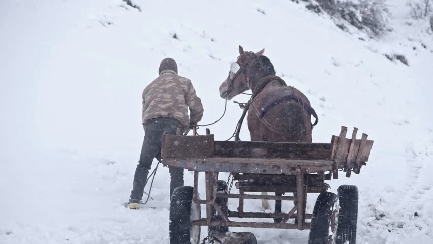 Two men walk in line with two horses pulling wooden carts through falling snow. White mountain landscape and cold winter atmosphere create a dramatic rural countryside scene.