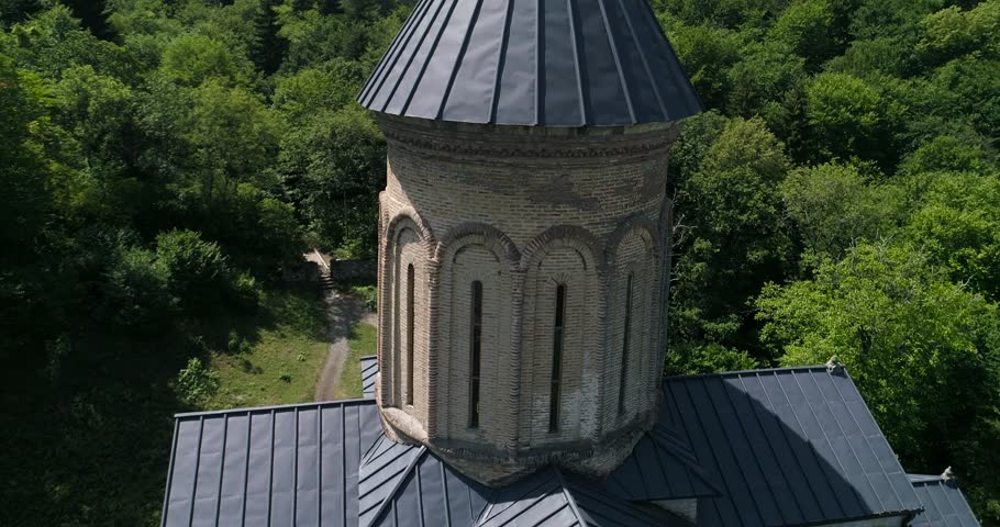 Drone close-up of a historic stone church tower with dark metal roof above green forest and trees.