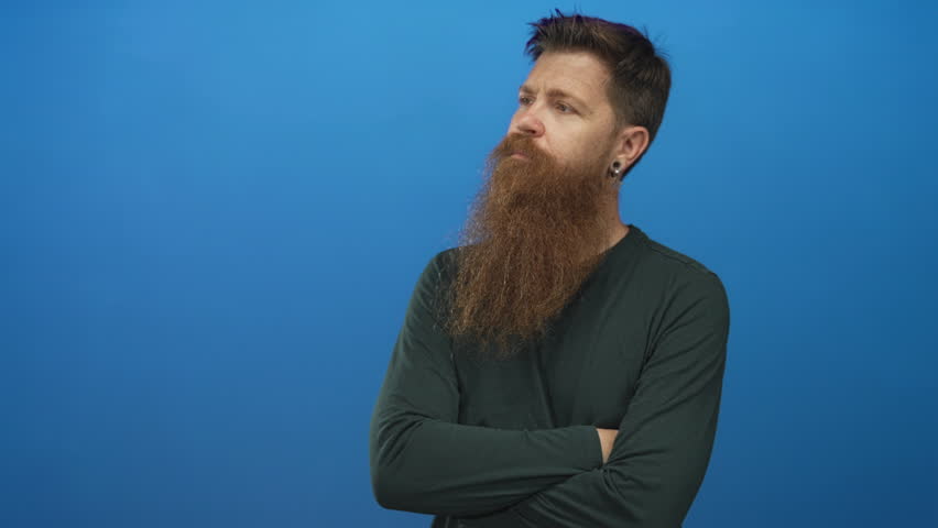 Young caucasian man with long red beard and earrings crosses arms in blue studio; contemplation calm focus.