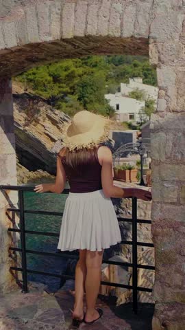 Woman in straw hat and white fluffy skirt standing in stone arch slow camera zoom reveals turquoise sea cliffs and small Petrovac fortress Montenegro