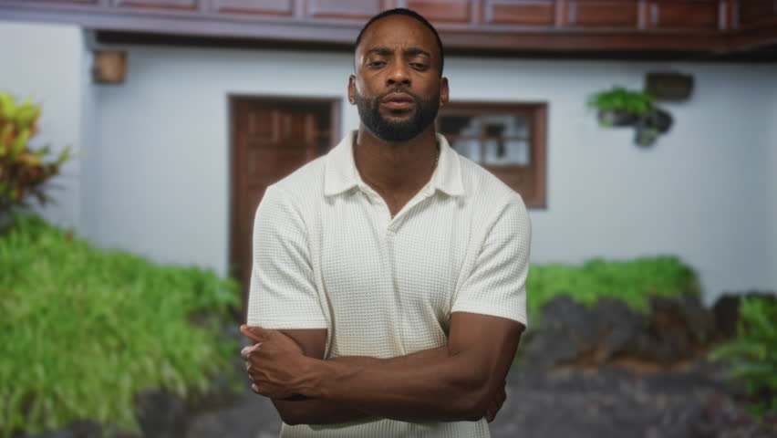 Young man with arms crossed at house entrance building, wearing white polo and visible forearms and beard; confidence.