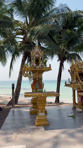 Traditional Thai spirit houses decorated with offerings on tropical beach in Thailand. Cultural shrine under palm trees by turquoise sea on island coastline.