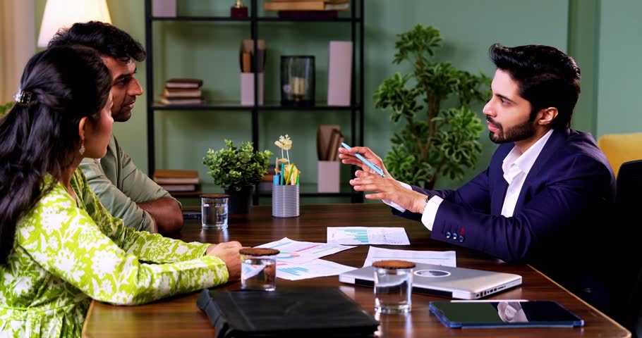 Indian couple meeting shaking hands with financial planner or ca in his office with laptop after reviewing growth reports and projections during serious investment discussion across table in office