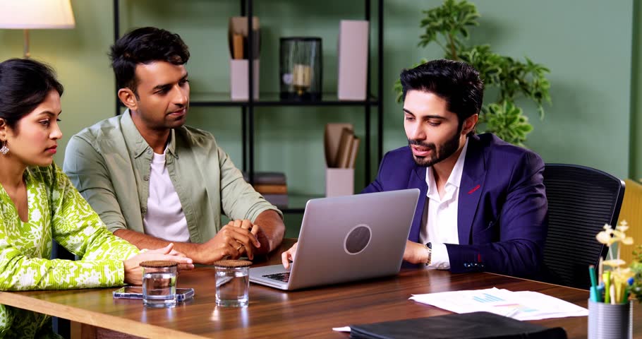 Indian couple visiting bank officer with laptop sitting across table during financial consultation discussing investment and real estate plans in modern corporate office interior setting