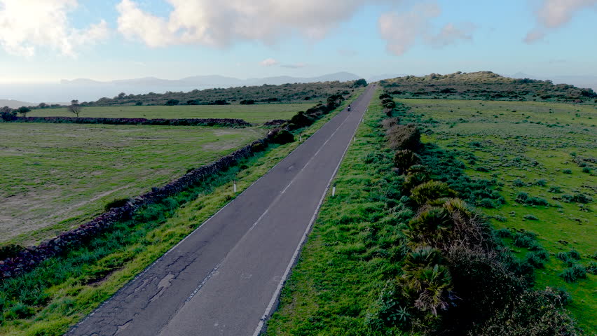 Aerial view of a classic motorcycle riding on a quiet country road during golden hour, approaching under a fixed drone camera and exiting the frame. 4K 60fps.