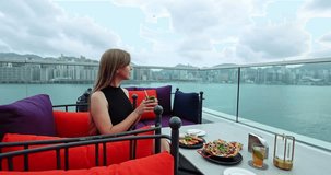 Woman sipping a refreshing cocktail and enjoying a meal at a luxurious rooftop restaurant overlooking a vibrant city skyline and tranquil harbor. Urban escape, modern lifestyle, luxury travel - Powered by Shutterstock - Get 15% off with code: PIKWIZARD15