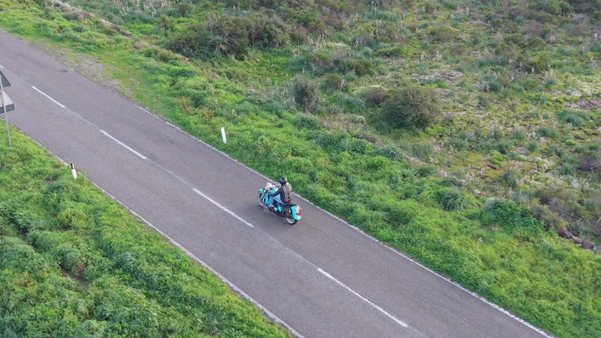 Aerial drone following a biker riding a classic motorcycle from a low 45Â° rear angle in slow motion on a country road. 4K 30fps footage.