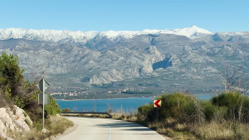 Handheld camera view from a moving vehicle, a curvy coastal road above the blue Adriatic Sea with the massive Velebit mountain range covered in snow. Bright winter sun and scenic Croatian coastline
