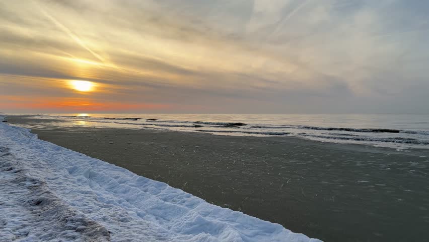 Golden sunset over the Baltic Sea in Latvia illuminating a snow covered shoreline and gentle winter waves, serene northern coastal landscape under dramatic evening sky.