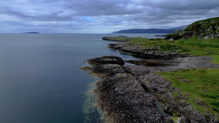 Aerial drone flight over rugged rocky coastline of Isle of Mull with green hills and calm sea. Dramatic Scottish island landscape under cloudy sky. Remote and wild Hebrides coastal scenery Scotland.