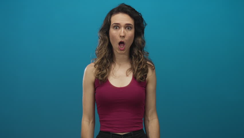 Woman in magenta tank top with open mouth and hands covering face in blue studio; surprise astonishment.