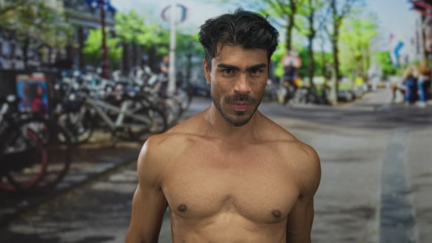 Man forming heart shape with hands over shirtless chest on sunlit street while smiling; love connection.