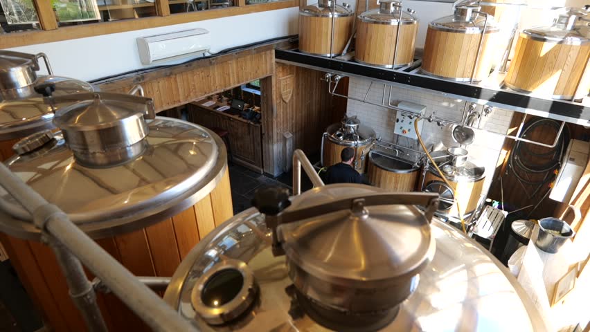 Slide left at three quarters angle on brewer inspecting beer brewery vats, interior with daylight