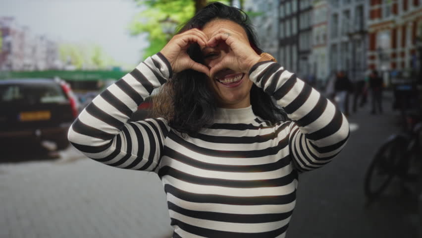 Woman wearing striped turtleneck makes heart gesture with hands in a busy city street, smiling and holding heart near face; joy love affection.