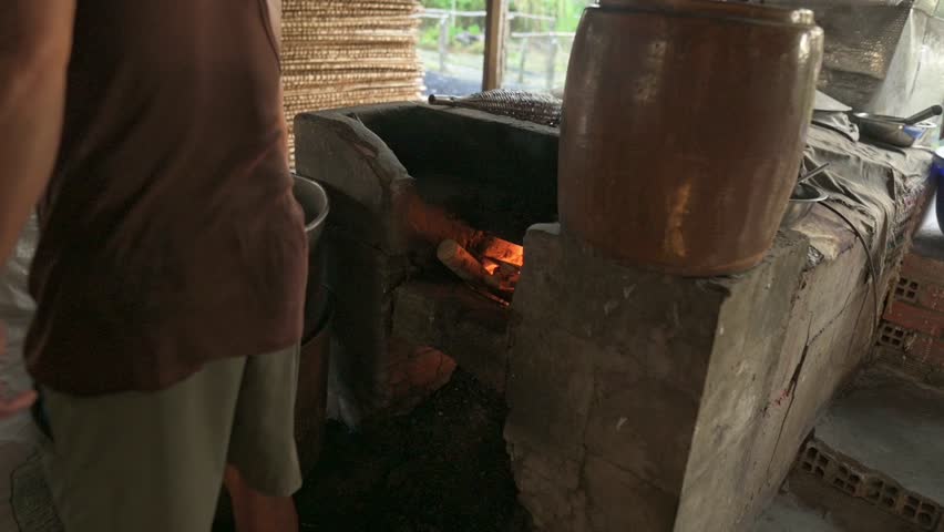 Local Cook Tending Wood Fire Stove, Adding Split Logs Into Clay Hearth, Glowing Embers Warming Ceramic Pot, Rustic Hut Kitchen Scene Showing Hands And Tools For Rice Noodle Prep