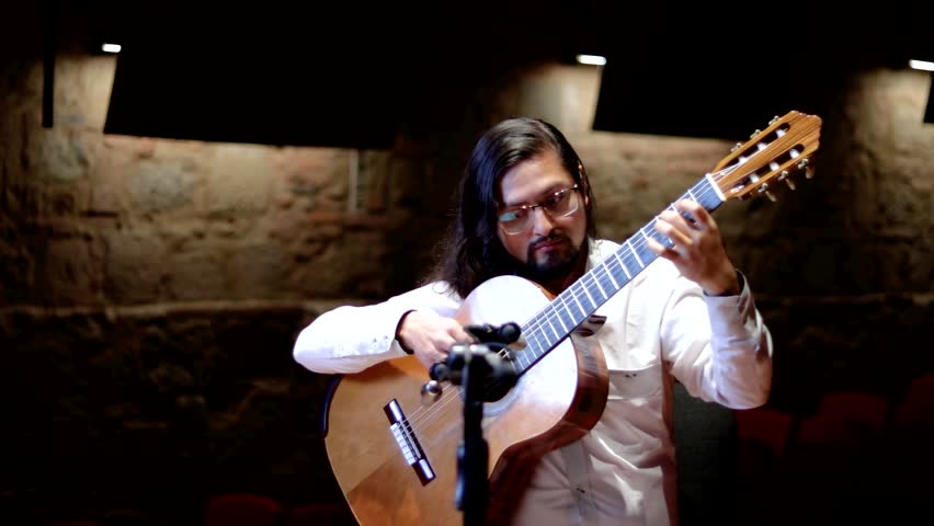 Horizontal shot of a male musician with long dark hair and glasses playing a classical acoustic guitar. He is wearing a white button-down shirt and is focused on his performance.