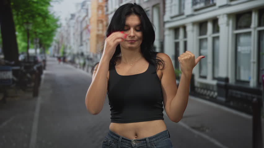 Woman in black tank top shows thumb to the side while standing on street with bare midriff exposed; indifference.