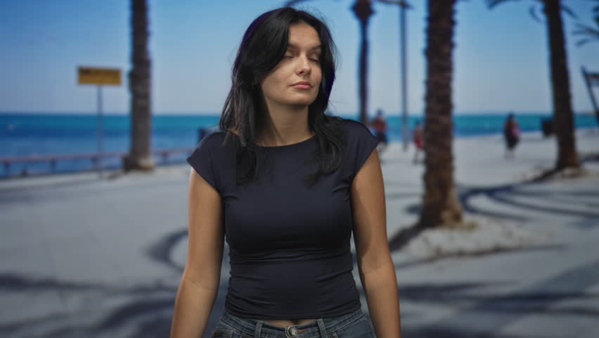 Woman wearing black top and jeans touches chest on coastal street under palm trees by the sea; calm reflection.