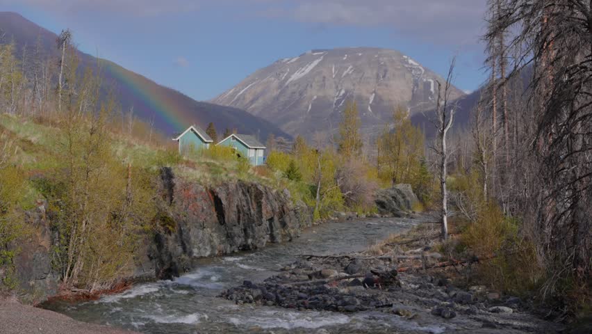 River flowing past cabins and mountains under a rainbow in Waterton Lakes National Park