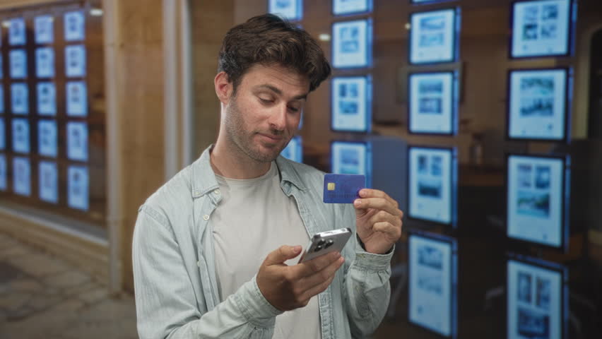 Man holding smartphone and creditcard in front of building with real estate listings; mobile payment confidence.