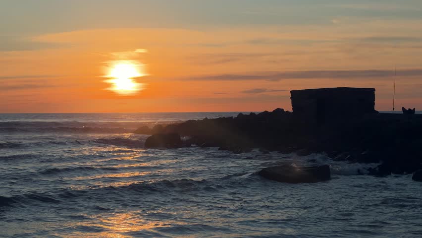 Fixed shot of sunset over the ocean with small stone breakwater silhouette and gentle waves in Barbate, Spain.