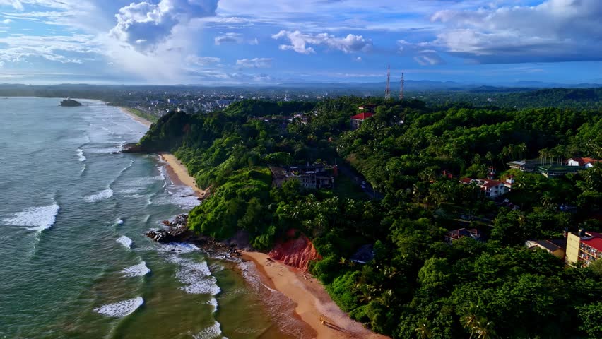 Aerial drone shot of Madiha Beach coastline with waves meeting sandy shore and lush green forest.