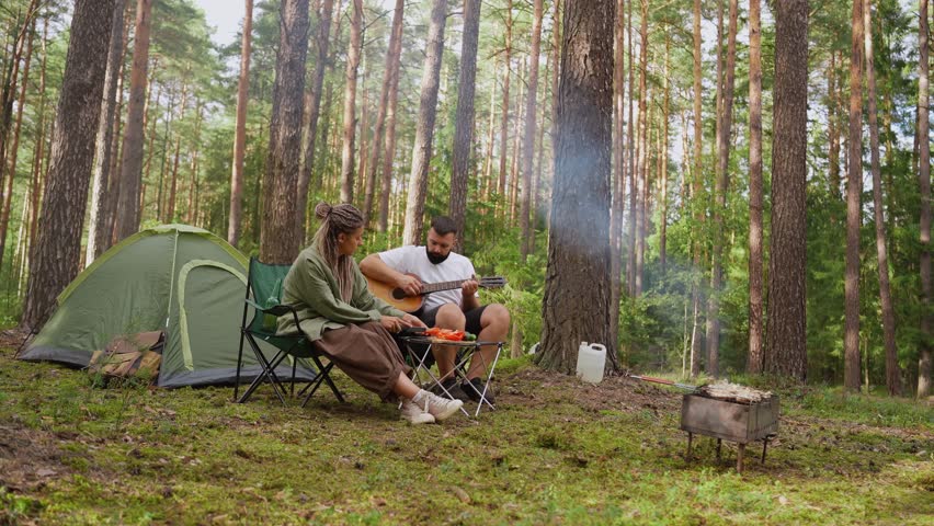 Young diverse couple enjoying a romantic picnic in the woods, with the man playing guitar while his girlfriend prepares food and meat grills on a barbecue next to their camping tent