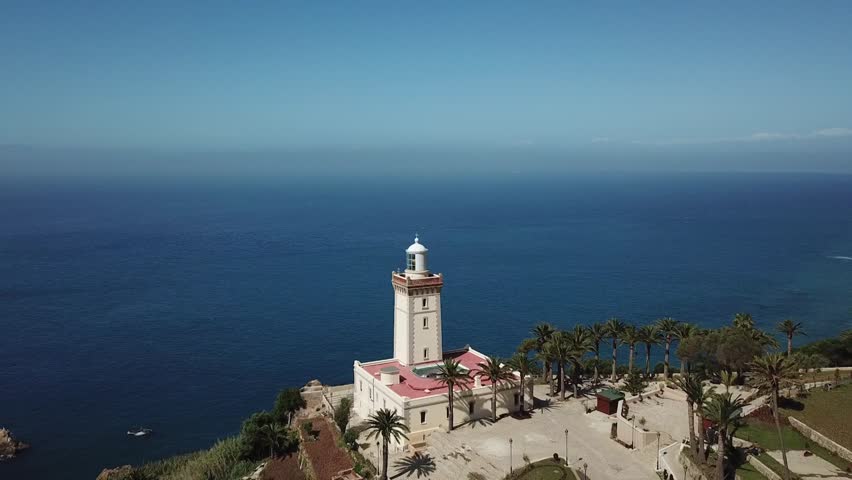 Majestic drone shot of the historic Cap Spartel lighthouse. Highlights the traditional architecture, palm trees, and the vast blue Atlantic horizon at the entrance of the Strait of Gibraltar.